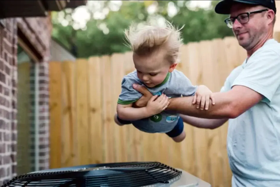 Mike Watson lifting his son next to an outdoor AC condenser install, a real moment at a Northern Kentucky HVAC shop